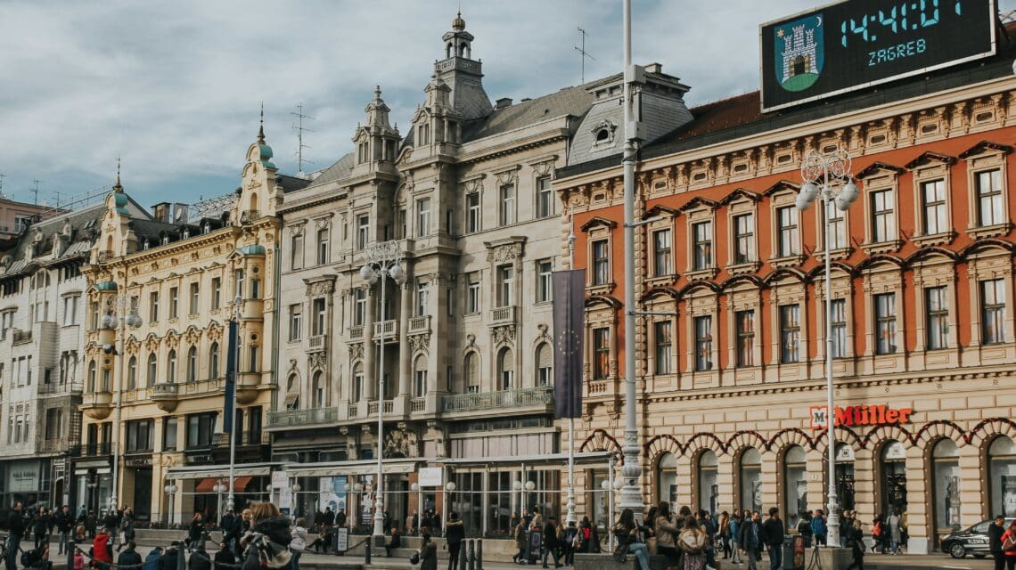 Historic European square with ornate pastel buildings under a cloudy sky. A digital clock shows 14:41:07, as people and pigeons fill the plaza.