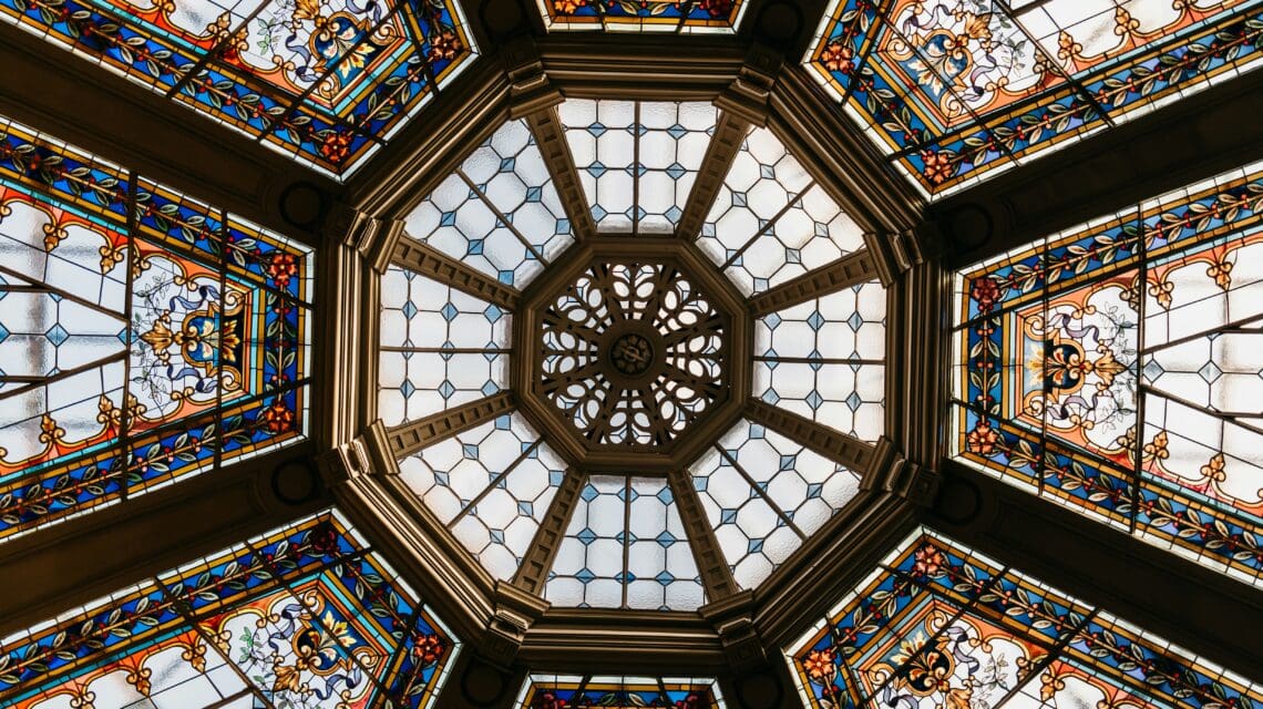 Ornate stained glass ceiling with a hexagonal center. Features intricate patterns in blue, orange, and gold. Elegant and historic architectural design.
