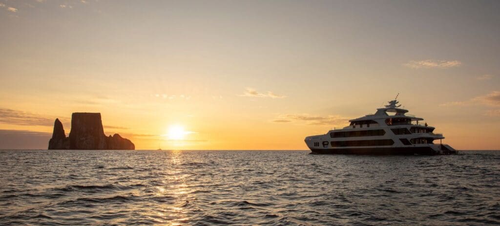 Sunset over calm ocean with a large yacht on the right and rocky outcrop on the left. Warm colors of the sky create a tranquil mood.