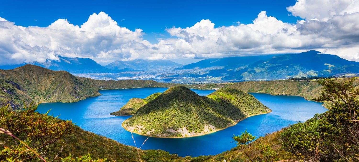 A serene lake with a lush, green island is surrounded by mountains under a clear blue sky with fluffy clouds. The scene is calm and picturesque.