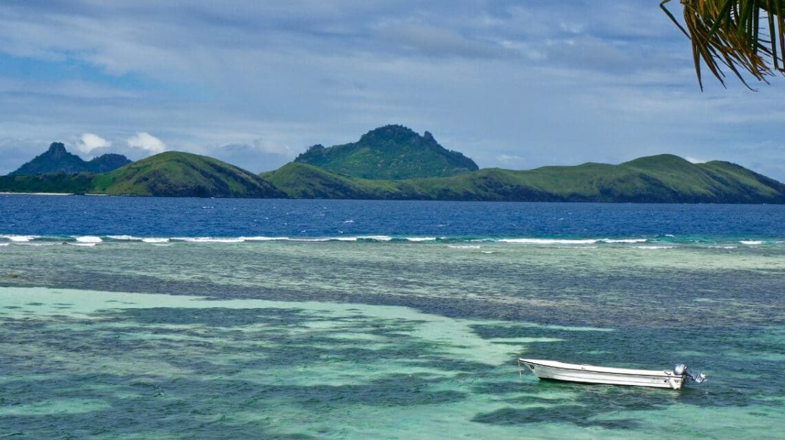 A serene coastal scene features a small boat on turquoise waters near coral reefs, with lush green islands and a cloudy sky in the background. Relaxing ambiance.