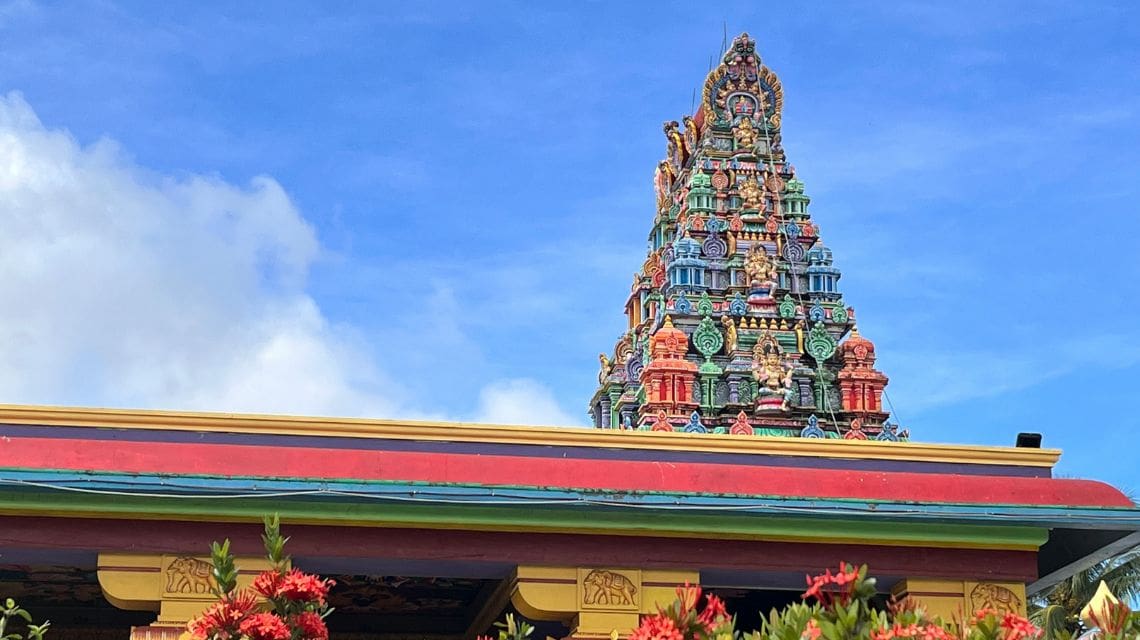 Colorful temple roof and ornate tower with intricate sculptures under a bright blue sky, surrounded by red flowers, conveying a vibrant, serene atmosphere.