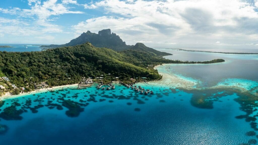 Aerial view of a tropical island with lush green mountains and turquoise waters. Overwater bungalows dot the coastline under a partly cloudy sky.