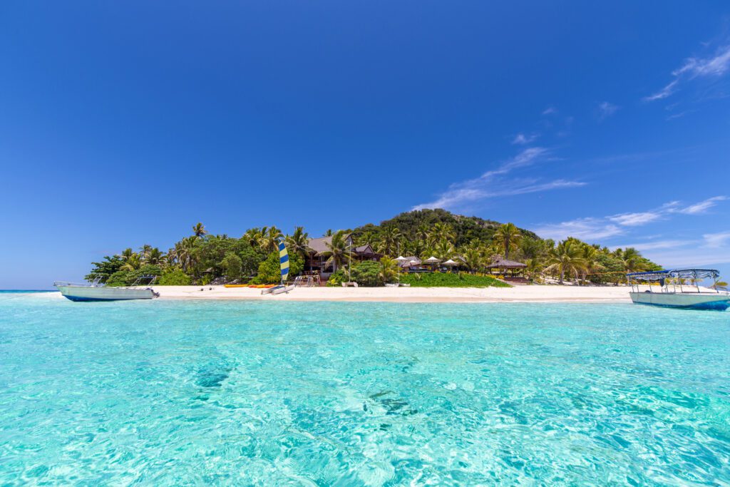 Tropical beach scene with clear turquoise water, white sand, and lush palm trees under a bright blue sky. Two boats sit on the calm water.