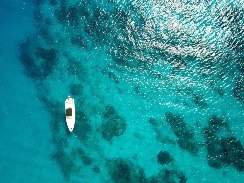Aerial view of a white boat on clear, turquoise water. Sunlight sparkles on the surface, casting shadows on coral reefs below, creating a serene scene.