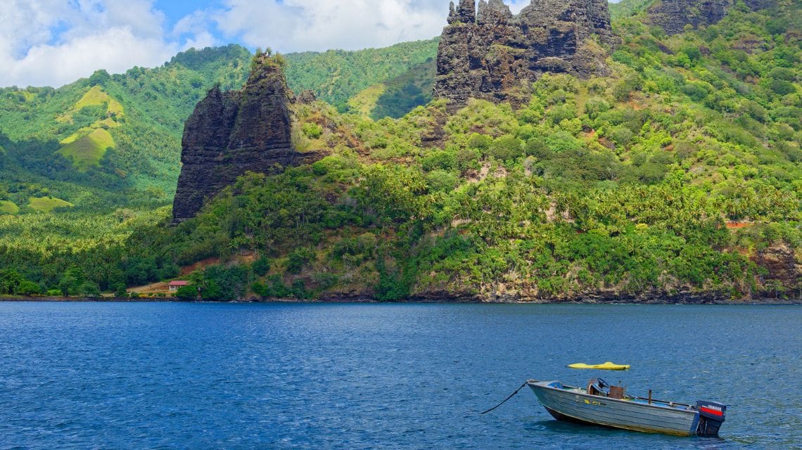 A small boat floats on calm blue water, with lush green hills and dramatic rock formations in the background. The scene evokes tranquility and natural beauty.