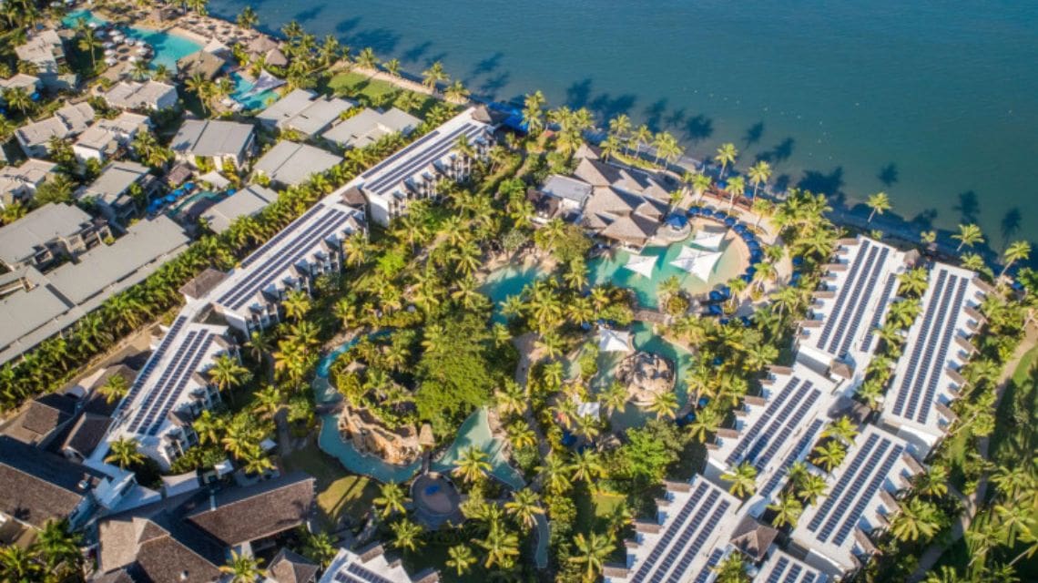 Aerial view of a tropical resort surrounded by lush palm trees and winding pools, next to a serene waterfront. Bright, sunny atmosphere.