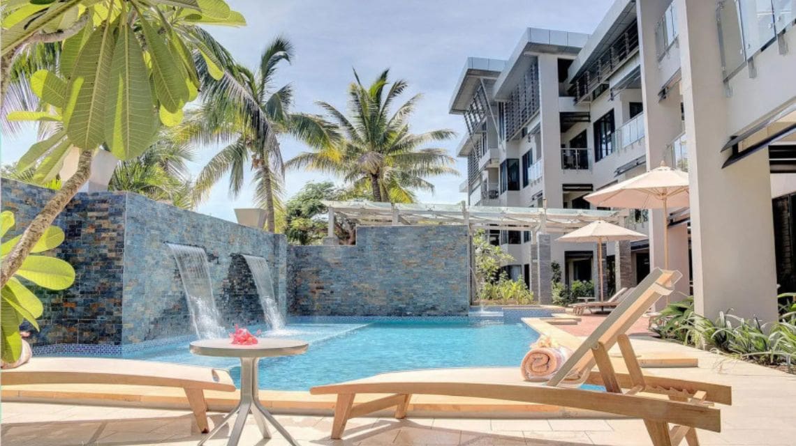 Sunny poolside scene at a resort with two waterfalls, palm trees, and modern buildings. Relaxed vibe with sun loungers and a small table with flowers.