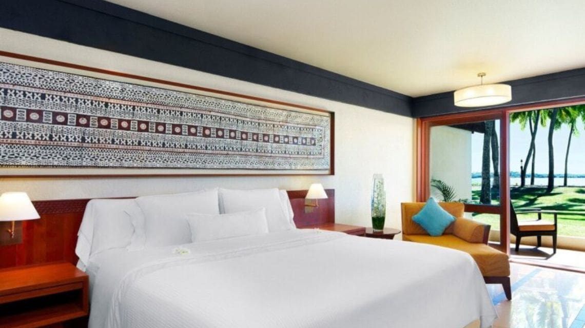A serene hotel room with a large white bed, abstract wall art, warm wooden accents, and a glass door opening to a sunny garden view with palm trees.