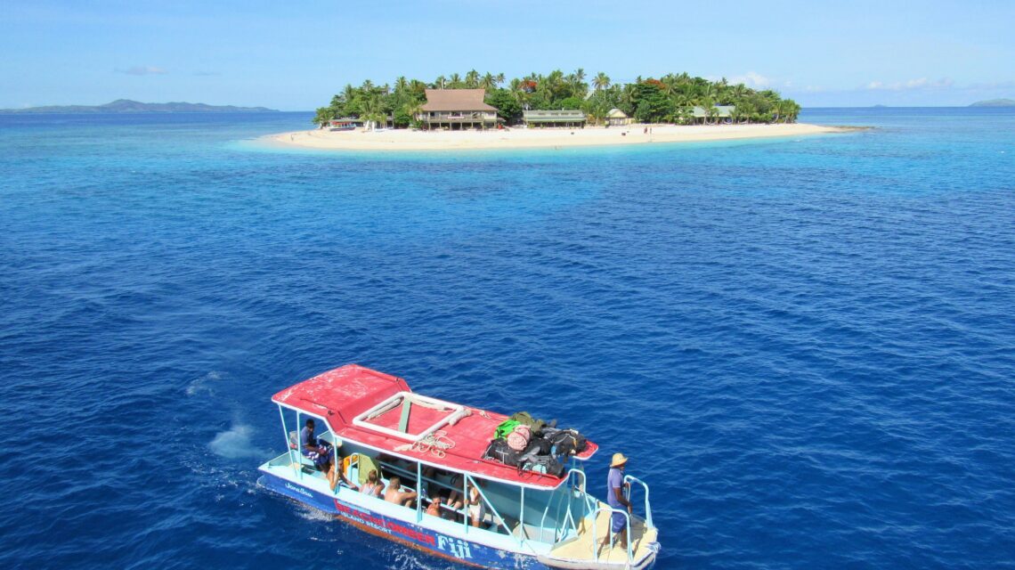 A small boat with people approaches a tropical island fringed with palm trees and white sand, under a clear blue sky, conveying a sense of adventure.