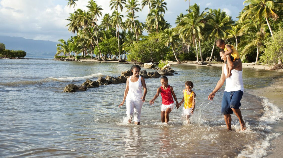 A happy family of five walks through shallow ocean waves at a tropical beach lined with palm trees, enjoying a sunny day in a joyful and relaxed atmosphere.