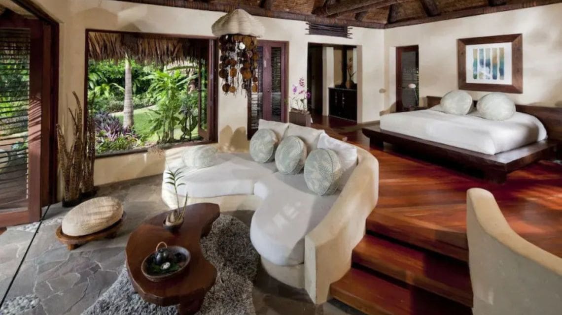 Luxurious tropical bungalow interior featuring a cozy white couch and wooden coffee table on a stone floor, large bed on a wooden platform, and lush garden view through open windows, conveying tranquility and warmth.