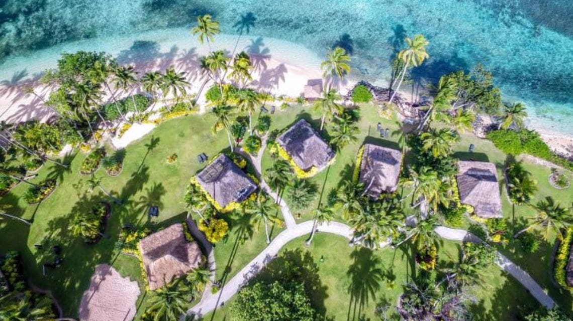 Aerial view of a tropical beach resort with straw-roofed huts surrounded by lush green palm trees, adjacent to turquoise ocean water and sandy shore.