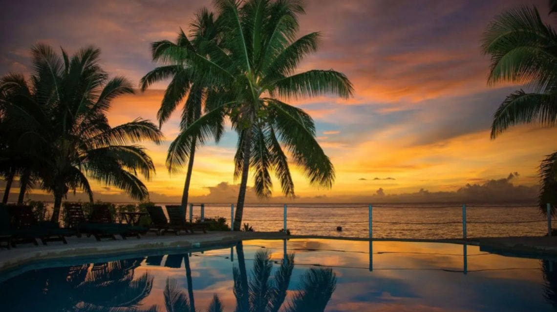 Tropical sunset with vibrant orange and purple hues over a calm ocean. Silhouetted palm trees and a still pool reflect the serene sky, evoking tranquility.