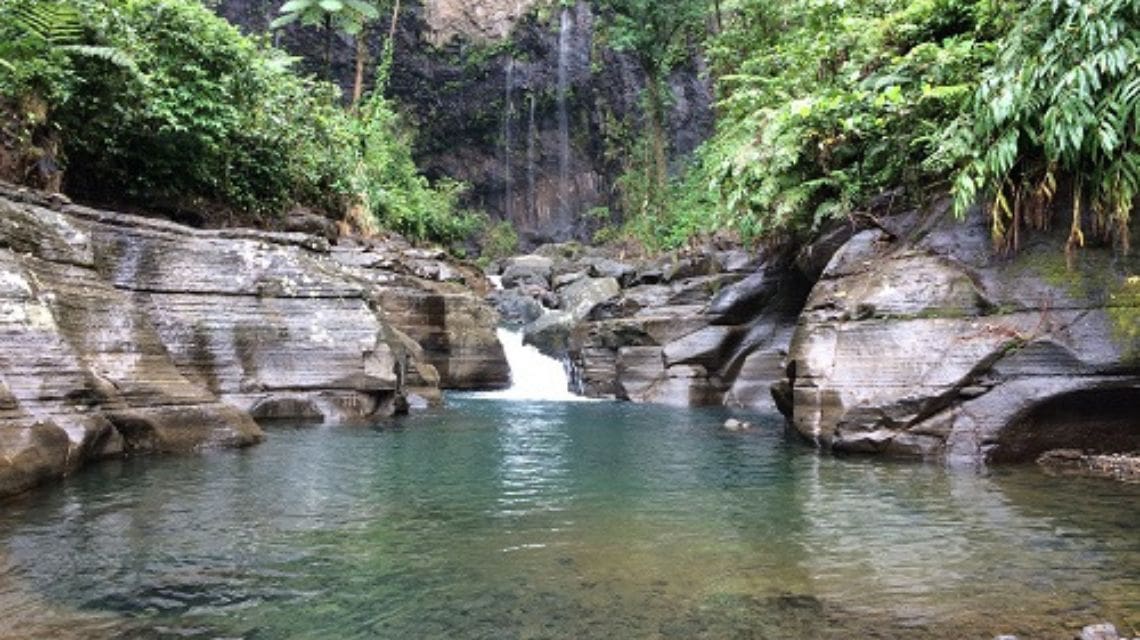 A serene waterfall cascades between rocky cliffs into a clear, emerald pool surrounded by lush green foliage, evoking tranquility and natural beauty.