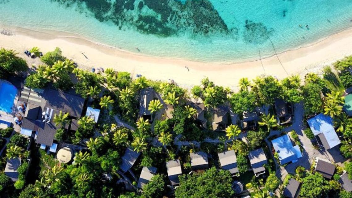Aerial view of a tropical beach with clear turquoise waters, beige sand, and lush green palm trees surrounding small villas, evoking serenity.