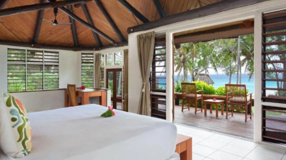 Tropical bedroom with wooden ceiling and open-air design, revealing a lush garden and ocean view. Cozy seating outside, conveying relaxation and tranquility.