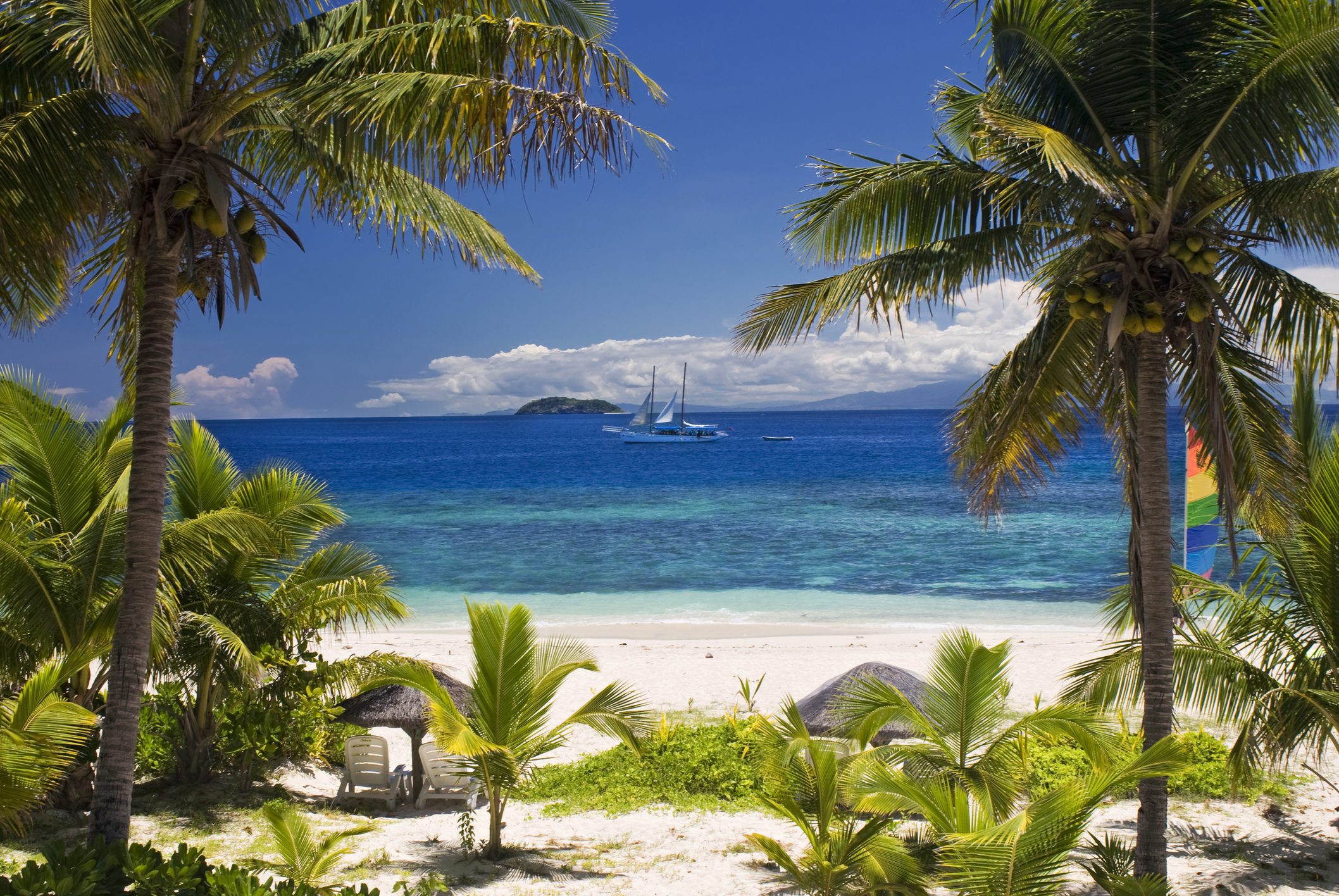 Tropical beach scene with palm trees framing a white sandy shore. A sailboat glides on the clear blue sea, and distant islands rest under a bright sky.