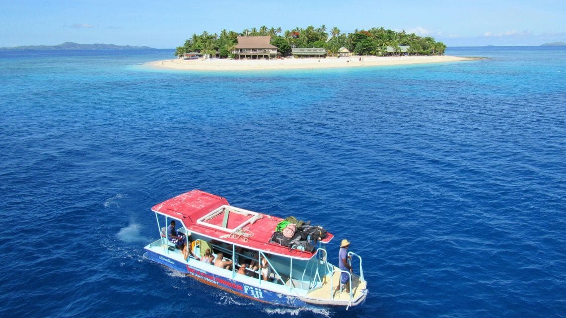 A small boat with passengers approaches a tropical island, surrounded by bright blue ocean. The island features a sandy beach and lush greenery.