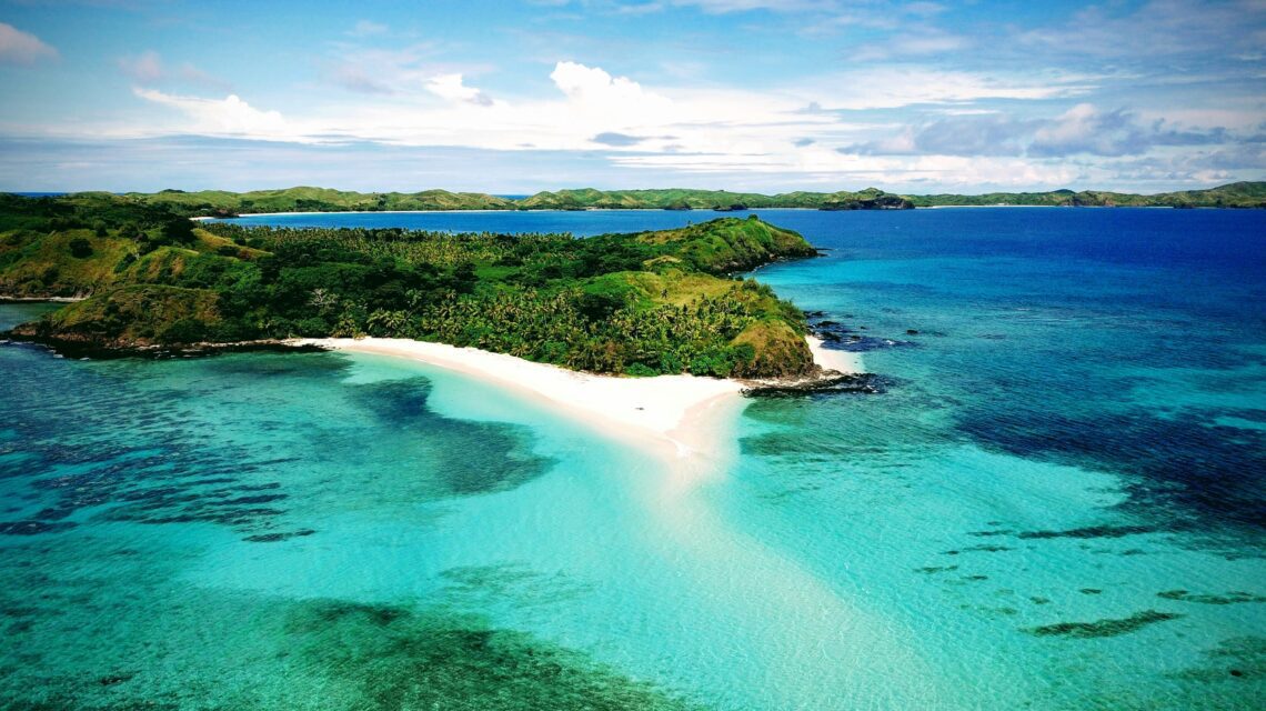 Aerial view of a tropical island with lush greenery and a pristine white sand beach surrounded by turquoise water, under a bright blue sky.