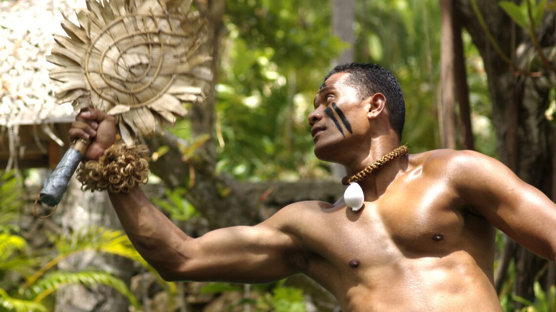 A man with dark painted stripes on his face performs a traditional dance in a lush tropical setting, holding a decorative fan. He appears focused and energetic.