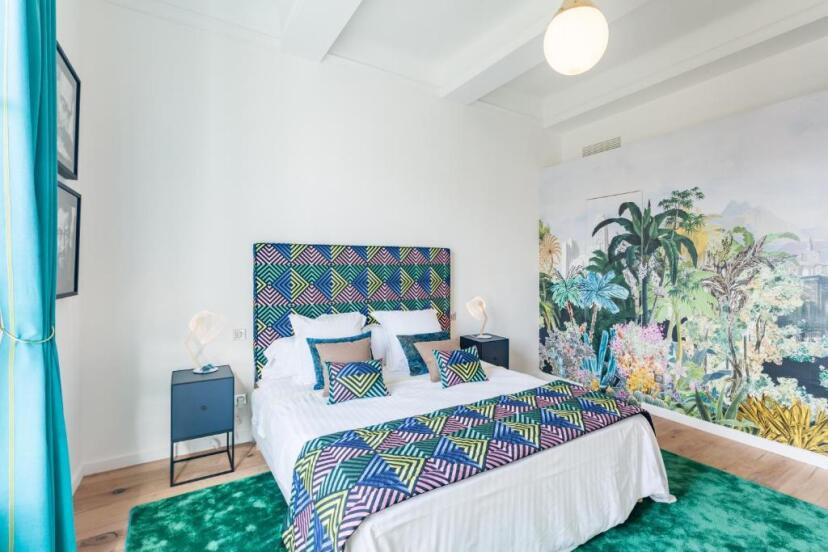 Bright bedroom with a colorful geometric headboard and vibrant pillows. A lush tropical mural covers one wall, creating a lively, fresh ambiance.