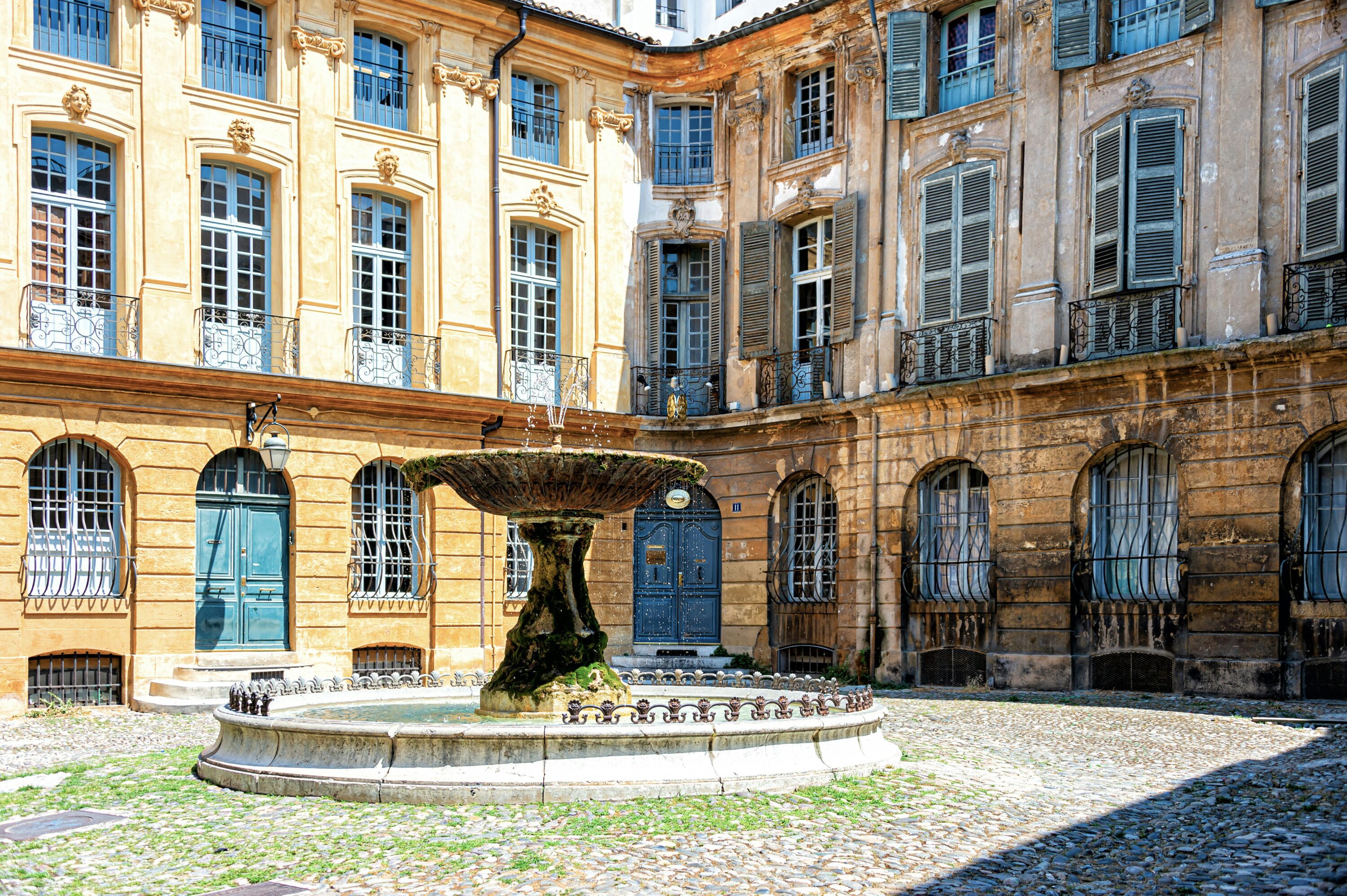 Historic courtyard with a weathered stone fountain at the center, surrounded by elegant, tall, ochre buildings with blue shutters and arched windows.