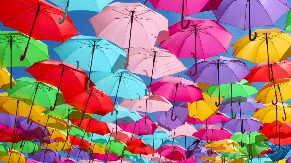 A vibrant canopy of colorful open umbrellas, including pink, red, green, and blue, hangs against a clear blue sky, creating a joyful and festive atmosphere.