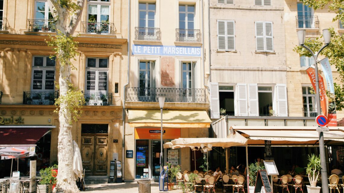 Street scene in a sunny European town with beige historic buildings, a café with outdoor seating under umbrellas, and leafy trees lining the street.