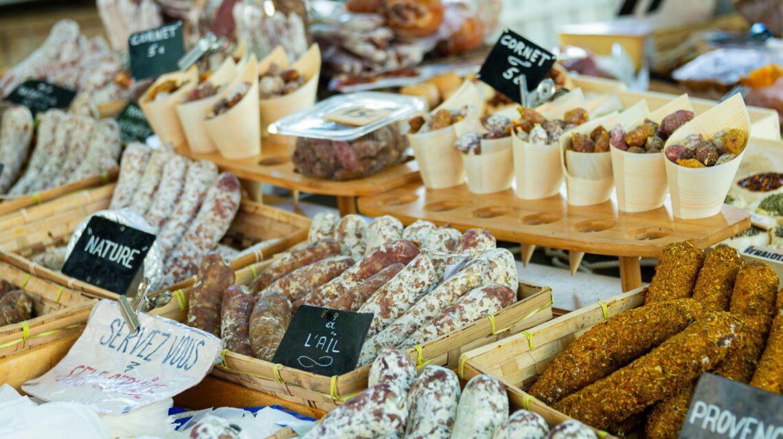 Assortment of cured sausages and meats with various seasonings displayed at a market. Baskets and signs convey a rustic and inviting atmosphere.