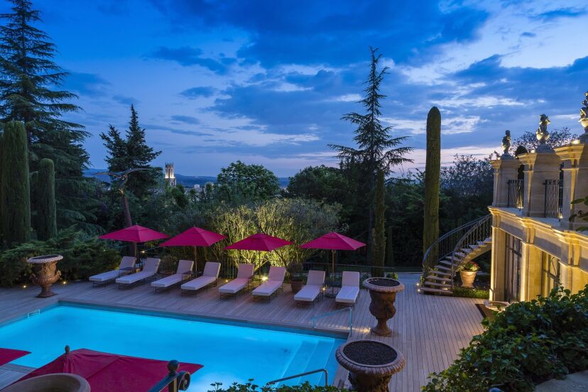 Luxurious poolside at twilight with seven red umbrellas and lounge chairs on a wooden deck, surrounded by lush trees and a lantern-lit stone staircase.