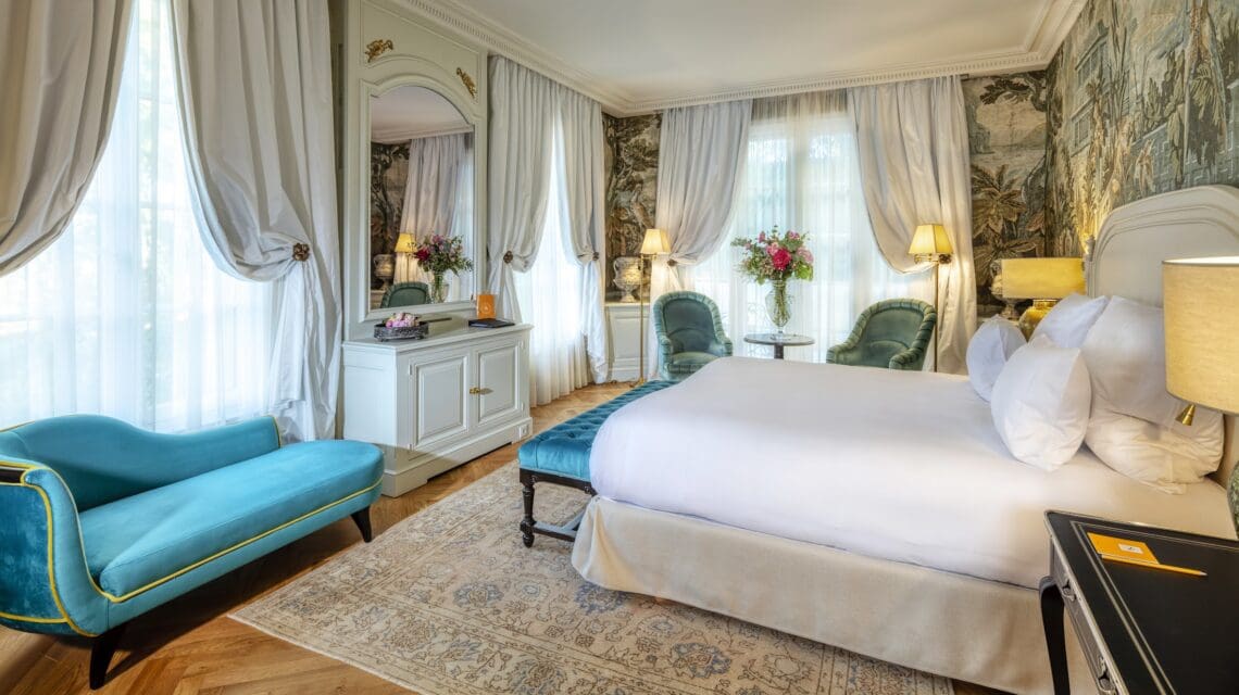 Elegant hotel room with a large bed, teal chaise lounge, and chairs. Soft lighting, floral wallpaper, and sheer curtains create a luxurious, serene atmosphere.