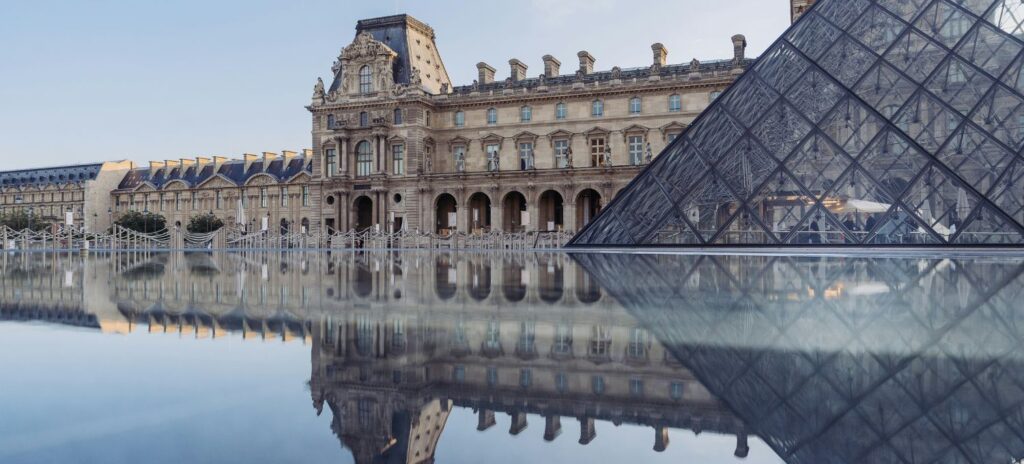 A glass pyramid and historic Louvre building are reflected in a calm water surface under a clear blue sky, capturing a serene and timeless scene.