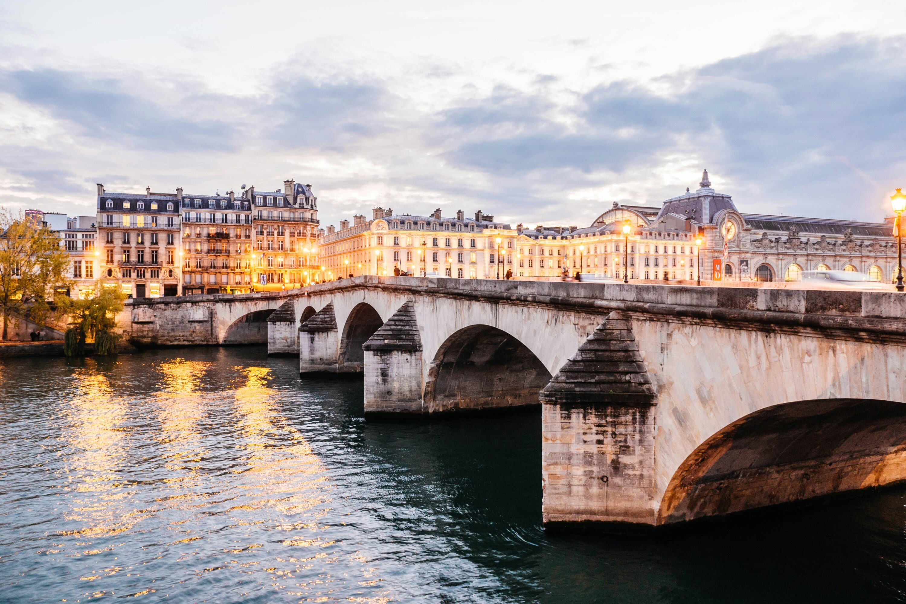 Scenic view of a historic stone bridge over the Seine River at dusk in Paris, with buildings illuminated by warm lights under a cloudy sky. Elegant and tranquil.