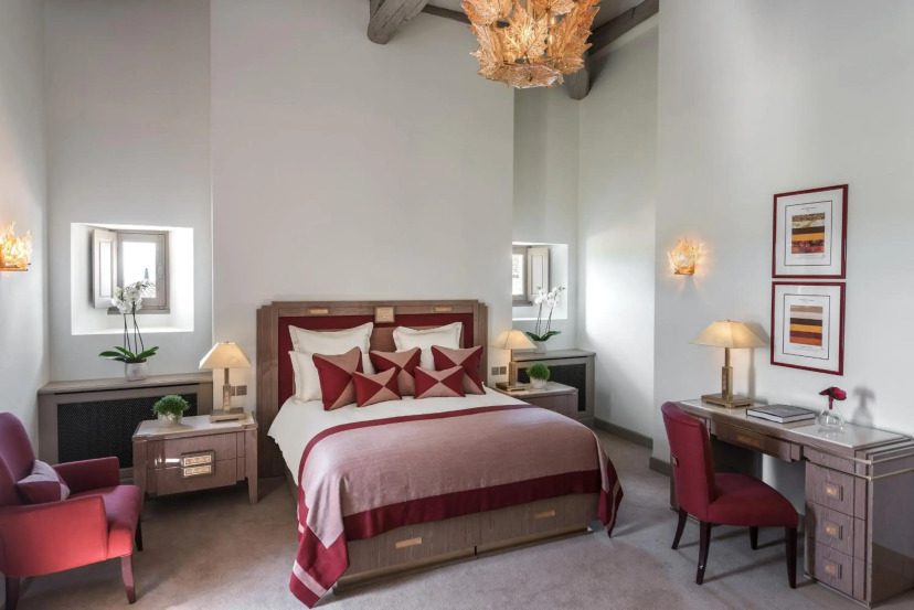 Elegant hotel room with a cozy double bed featuring red and white pillows, flanked by nightstands with lamps. A desk, two red chairs, and wall art add sophistication.
