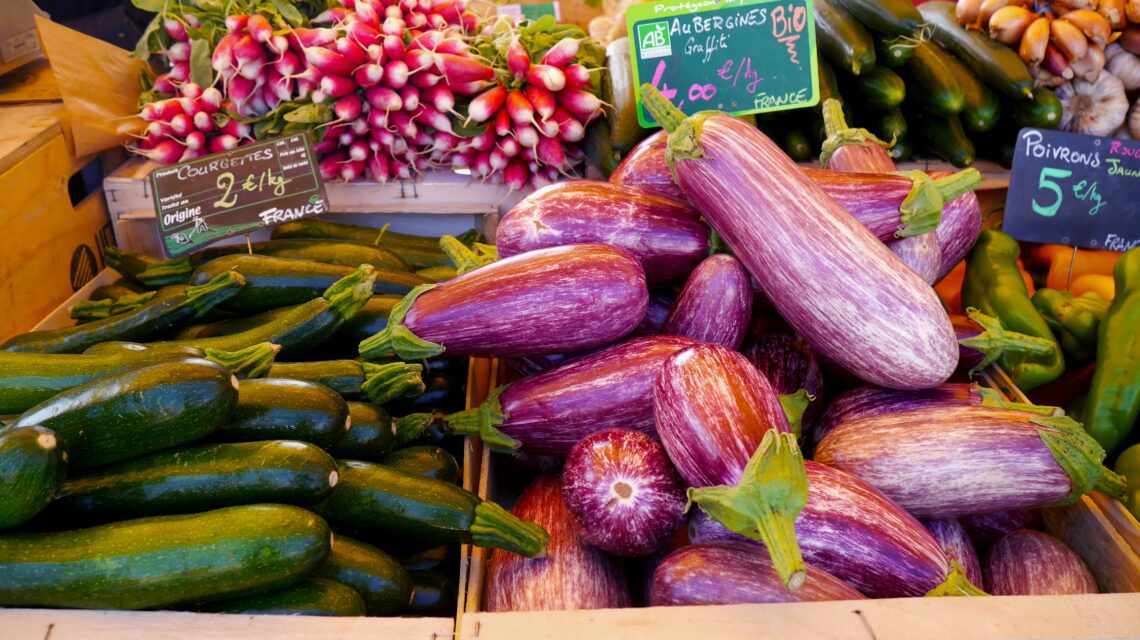 Vibrant market stall with striped eggplants, green zucchinis, and pink radishes. Colorful, fresh produce creates a lively, bustling atmosphere.