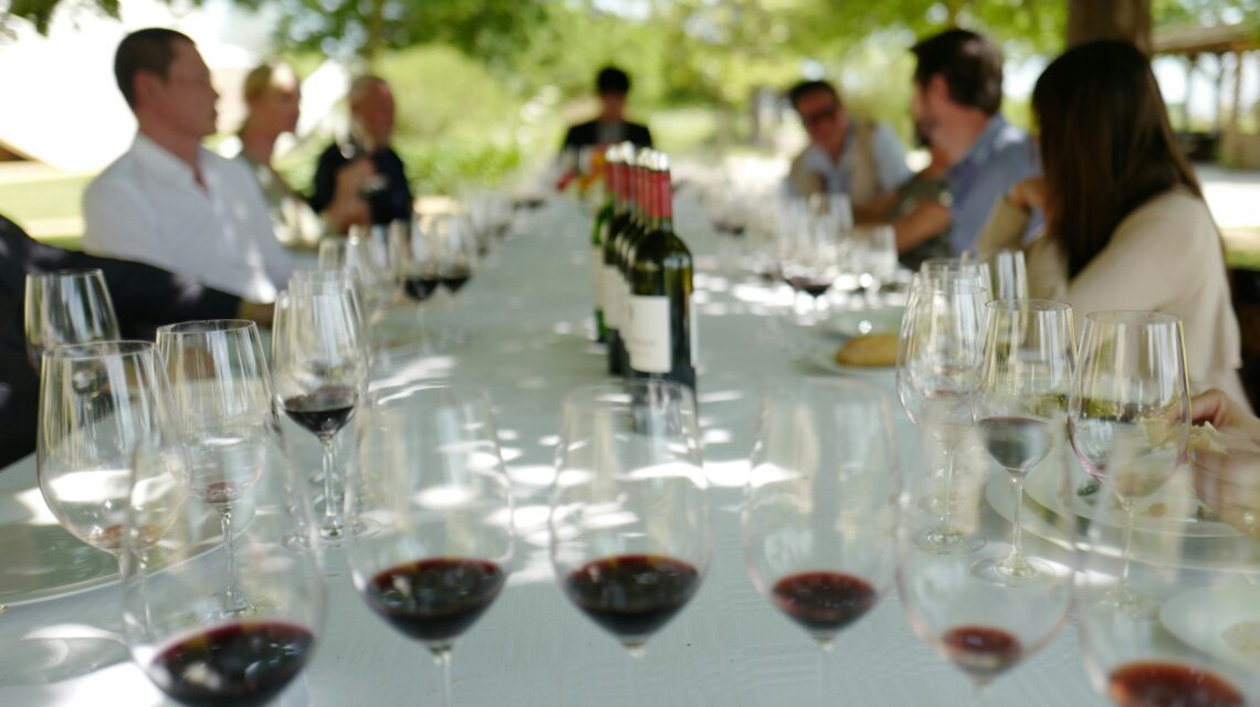 Outdoor wine tasting event under trees, featuring a long table with red wine glasses and bottles. Guests engage in conversation, creating a relaxed atmosphere.