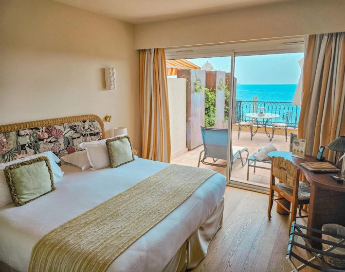 Cozy hotel room with a plush double bed, light decor, and sliding glass doors opening to a balcony with chairs overlooking a vibrant blue sea. Serene ambiance.