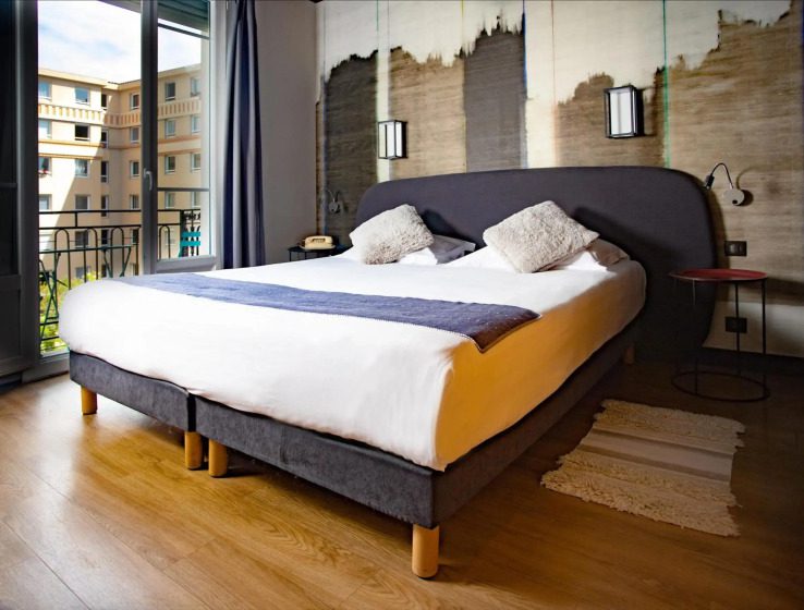 A modern bedroom featuring a large bed with white linens and gray decorative pillows. Soft lighting highlights an abstract mural above the headboard, creating a cozy, elegant atmosphere. The room has wooden floors, a bedside table, and a window view of buildings outside.