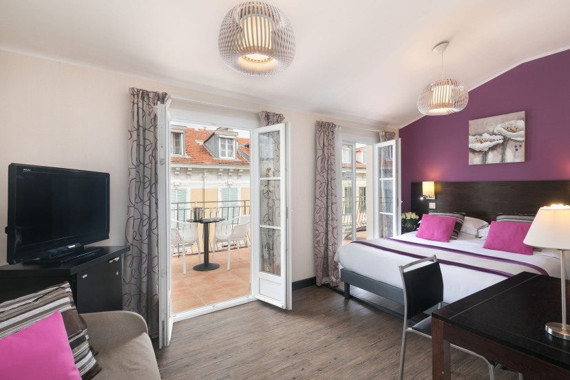 Bright hotel room with a purple accent wall, double bed with pink pillows, a balcony view, and contemporary decor. A TV and desk are on the side.