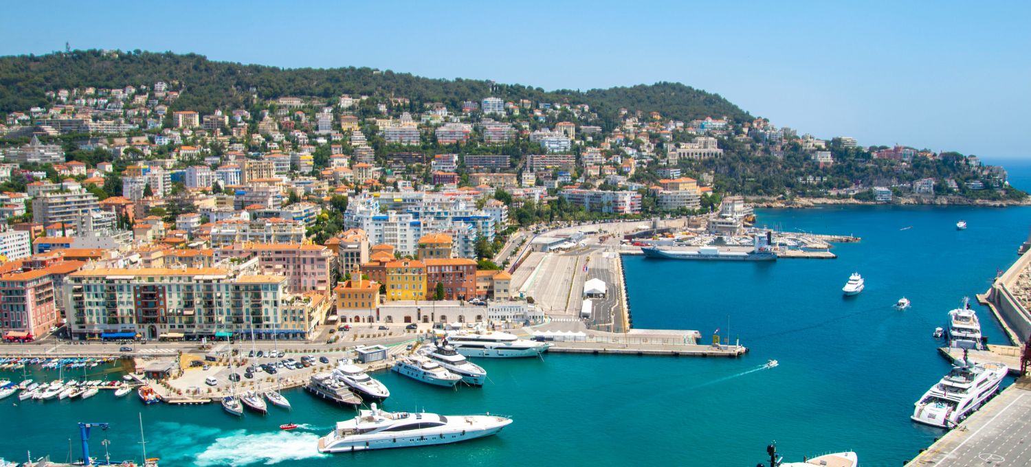 A vibrant marina with luxury yachts docked in clear blue water, bordered by colorful buildings and lush green hills. The scene is lively yet serene.