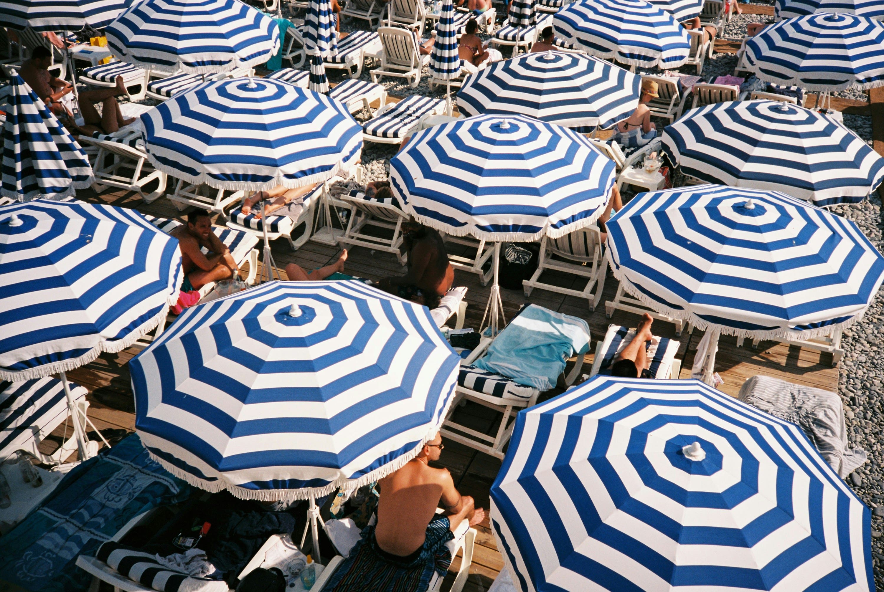 Aerial view of a crowded beach with blue and white striped umbrellas. Sunbathers relax on lounge chairs, creating a lively, summery atmosphere.