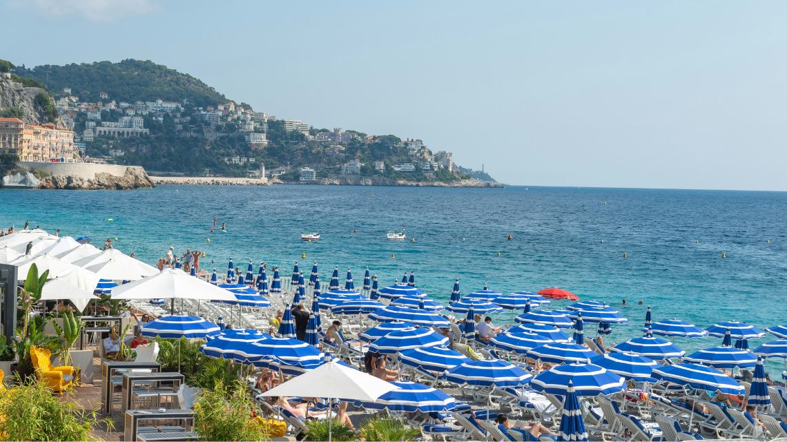 A sunny beach crowded with blue and white striped umbrellas and loungers. Tourists relax by the turquoise sea, with hills and coastal buildings in the background.