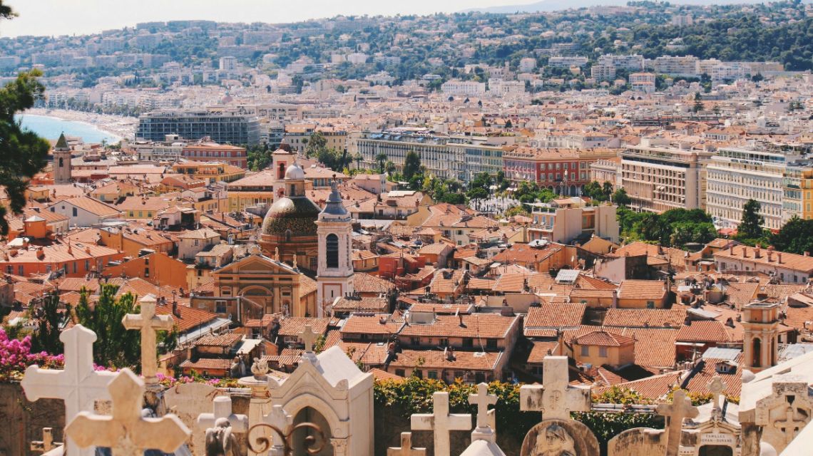 A panoramic view of Nice, France, featuring terracotta rooftops, a prominent church dome, and a distant coastline under a clear sky. The scene is vibrant and historic.
