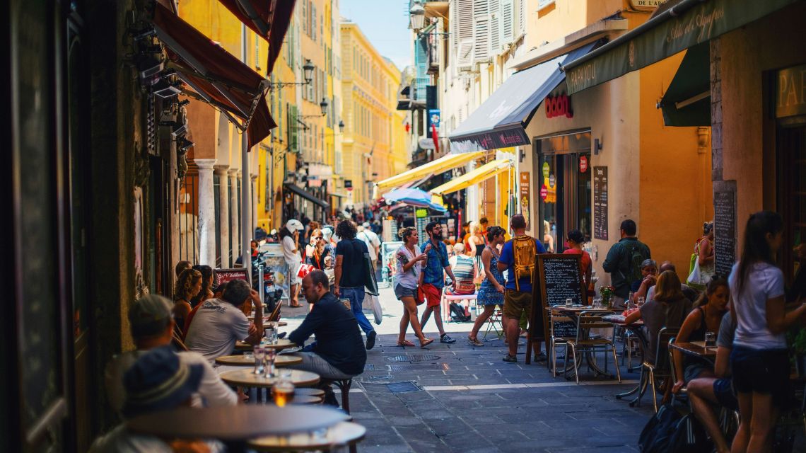 A bustling European street scene with outdoor cafes, people walking, and vibrant yellow buildings. Sunlit and lively, conveying a warm, cheerful atmosphere.
