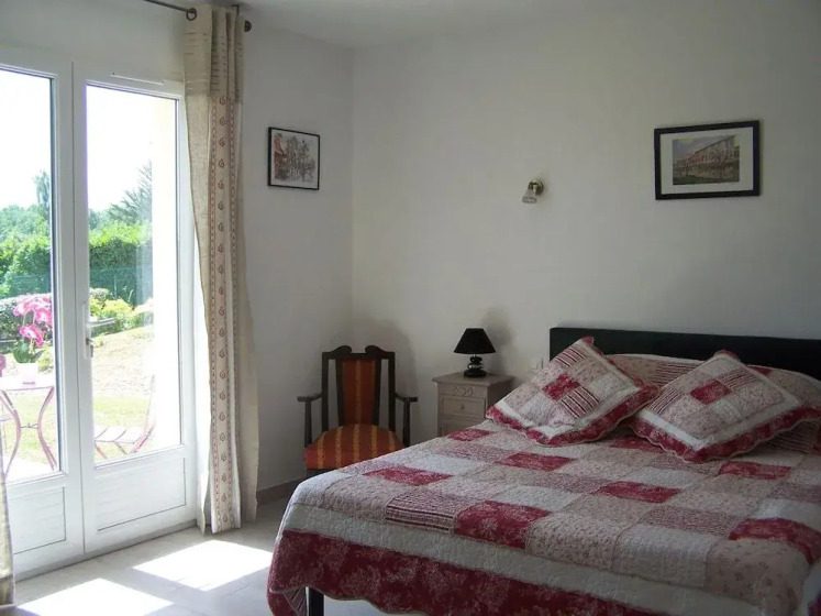 A bright bedroom features a bed with a red and white patchwork quilt and cushions, an ornate chair, and a side table. French doors and framed art add charm.