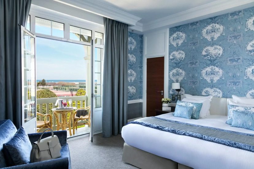 Elegant hotel room with blue patterned wallpaper, a plush bed, and chic decor. Open doors reveal a balcony with a scenic ocean view. Relaxing ambiance.