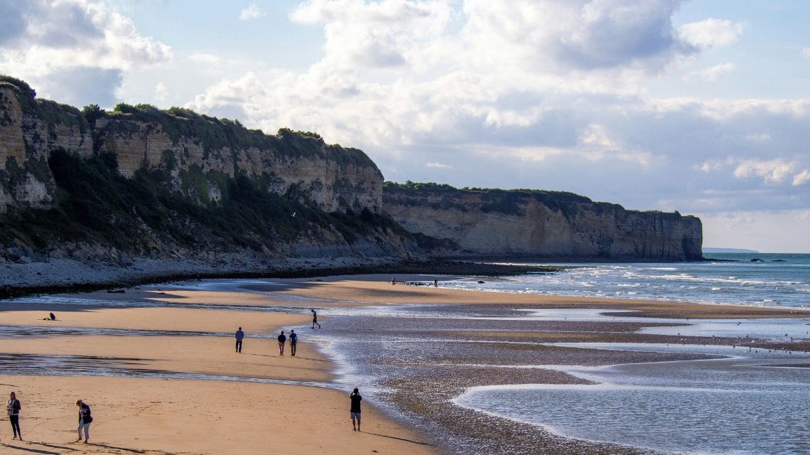 A scenic view of a coastal beach with dramatic cliffs under a cloudy sky. People stroll along the sand, evoking a tranquil and peaceful atmosphere.