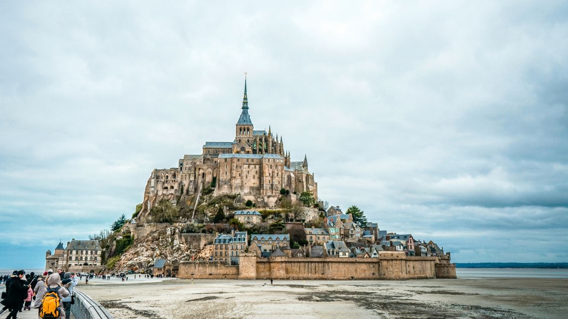 A grand medieval abbey and village rise on Mont Saint-Michel, surrounded by expansive sandy flats under a cloudy sky. Tourists walk along the causeway.
