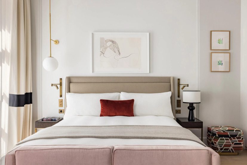 Modern bedroom with a plush bed featuring a red accent pillow, neutral tones, abstract wall art, a stylish hanging light, and elegant decor. Calm ambiance.